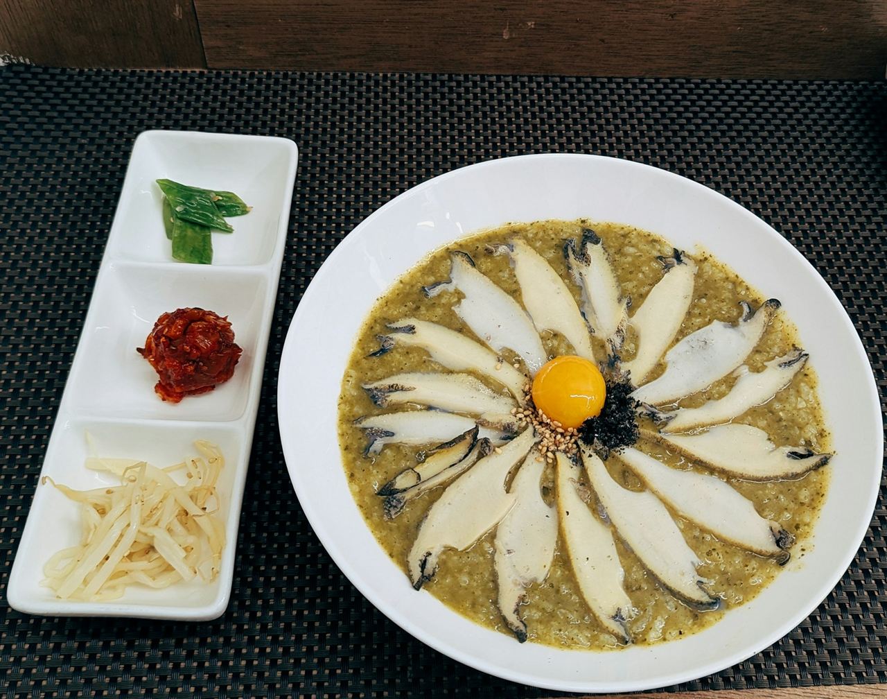 A wide bowl of green abalone porridge topped with sliced abalone and an egg yolk, with small side dishes