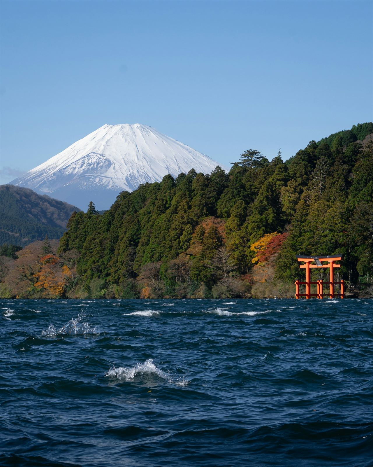 Snow-capped Mount Fuji above the forested shore of Lake Ashi, a red torii gate at the water’s edge