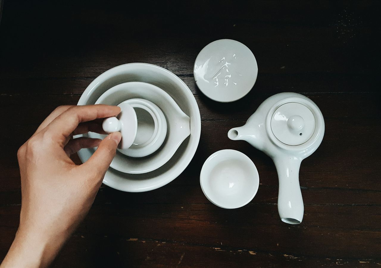 A white porcelain tea set on a dark wooden table, a hand lifting the lid of a bowl