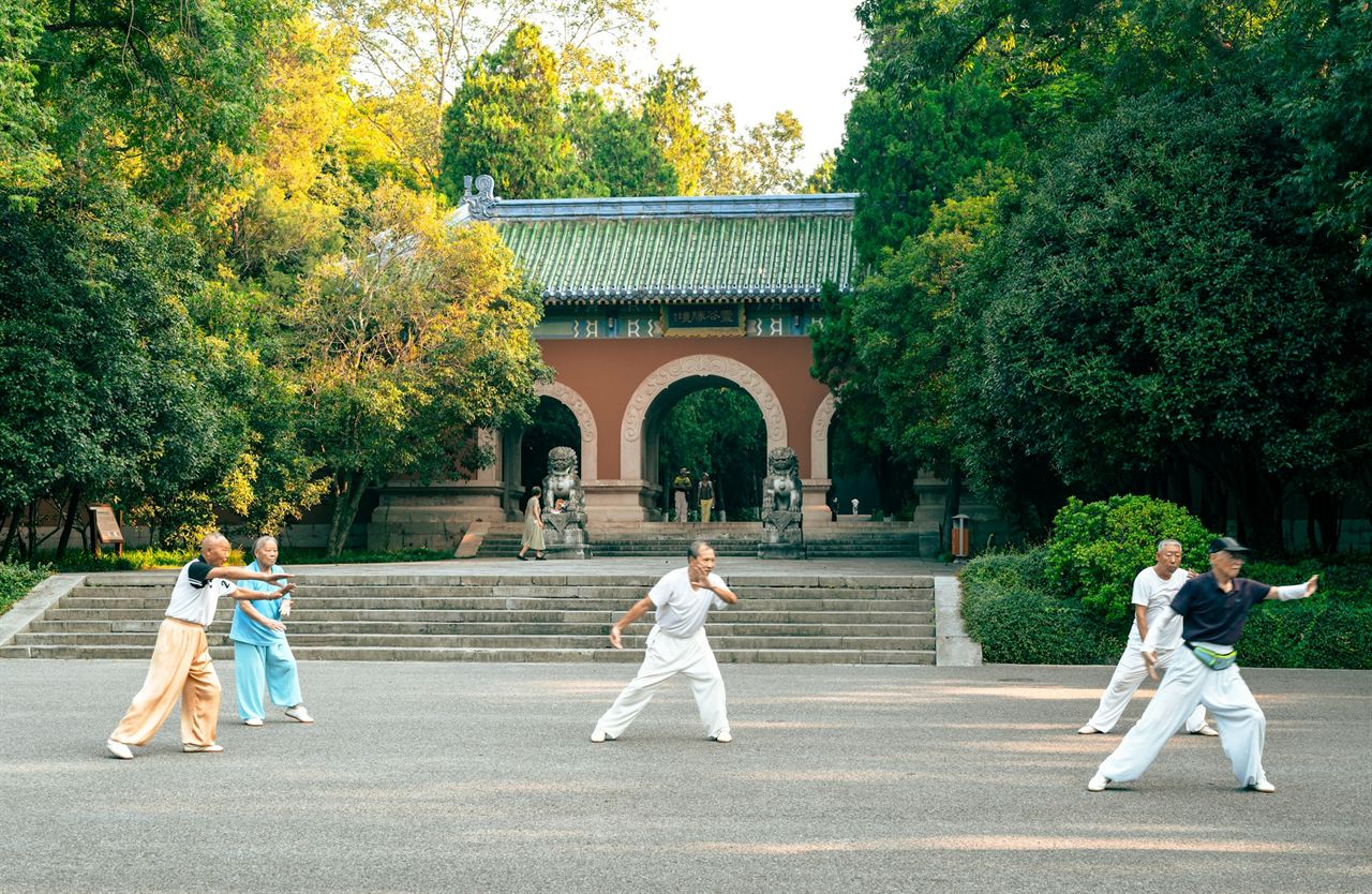 People in loose white clothes practicing tai chi on a sunny square in front of a temple gate