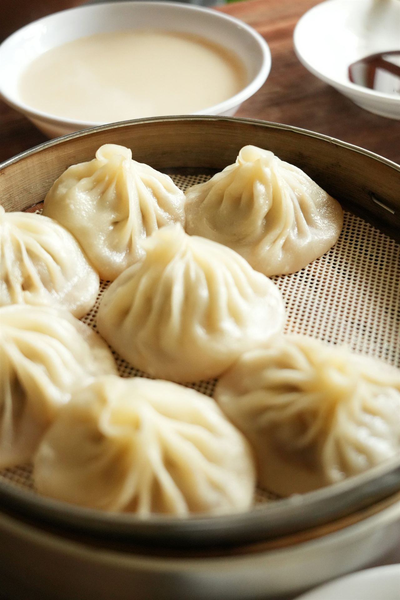 A bamboo steamer of soup dumplings beside a bowl of soy milk